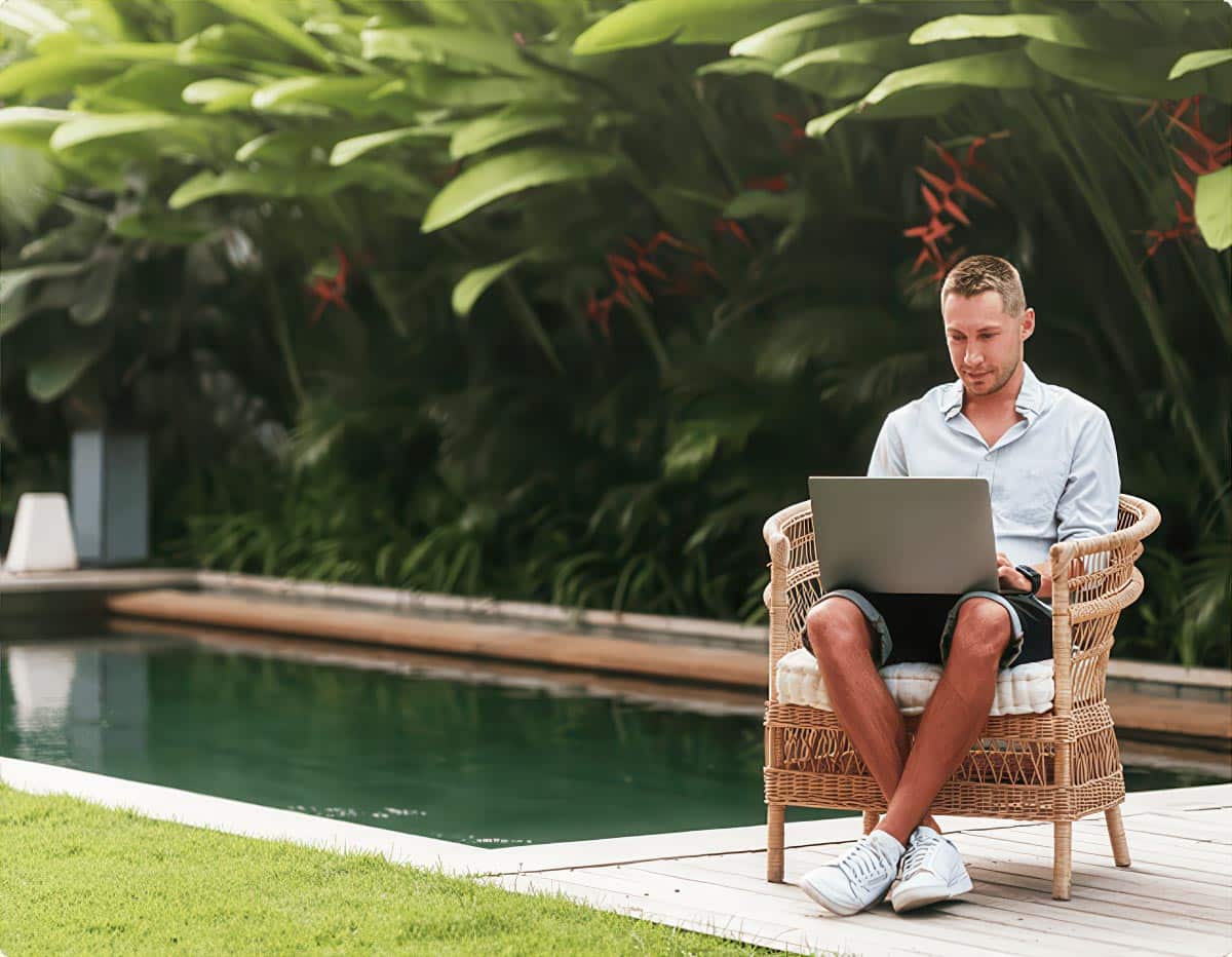 Man sitting poolside in wicker chair working on laptop